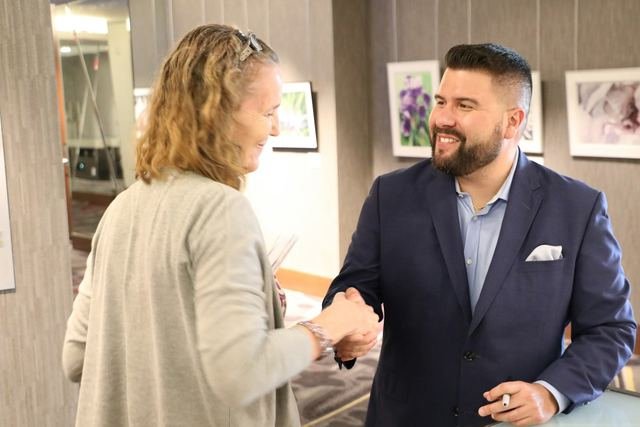 A smiling man in a navy suit shakes hands with a woman during a friendly meeting