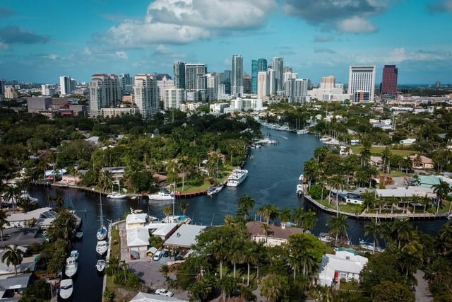 Aerial view of waterfront homes along palm-lined canals with boats at private docks and the Fort Lauderdale skyline behind